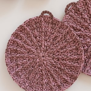 Pode incluir: Dois pegadores de panela de crochê em um esquema de cores rosa e marrom. Os pegadores de panela são redondos e têm um design texturizado.