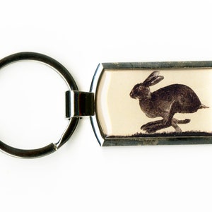 Puede incluir: Un llavero de plata con un anillo redondo y una etiqueta rectangular. La etiqueta tiene un fondo blanco con una imagen en blanco y negro de una liebre corriendo.