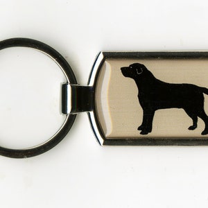 Puede incluir: Un llavero rectangular con un marco de metal plateado y una silueta de perro negro sobre un fondo beige. El llavero tiene un gran llavero circular adjunto al marco.
