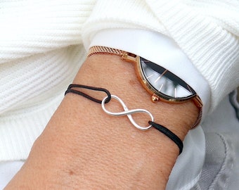 May include: A black string bracelet with a silver infinity charm. The bracelet is worn on a wrist with a rose gold watch.