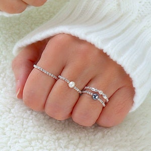 Könnte beinhalten: Eine Hand trägt vier silberne Ringe mit kleinen Perlen und einer Perle. Die Ringe sind auf zwei Fingern übereinander gelegt.