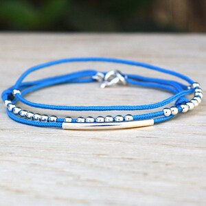 Op de afbeelding: Een blauw koordarmband met zilveren kralen en een zilveren staafbedel.