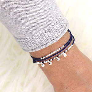 May include: A layered bracelet with navy blue cord and silver beads. The bracelet features a silver bar charm and dangling silver ball charms.