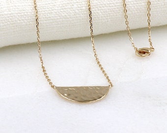 Può includere: Una collana color oro con un ciondolo a mezzaluna martellata. La delicata catena e il ciondolo sono presentati su una superficie bianca, creando un'estetica minimalista. La collana è un gioiello semplice ma elegante.
