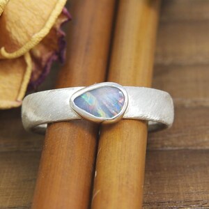 May include: A silver ring with a textured band and a teardrop-shaped opal gemstone set in the centre. The opal has iridescent colours of blue, green, and purple.