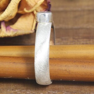 May include: A silver ring with a brushed finish and a round, flat top. The ring is resting on a wooden surface.