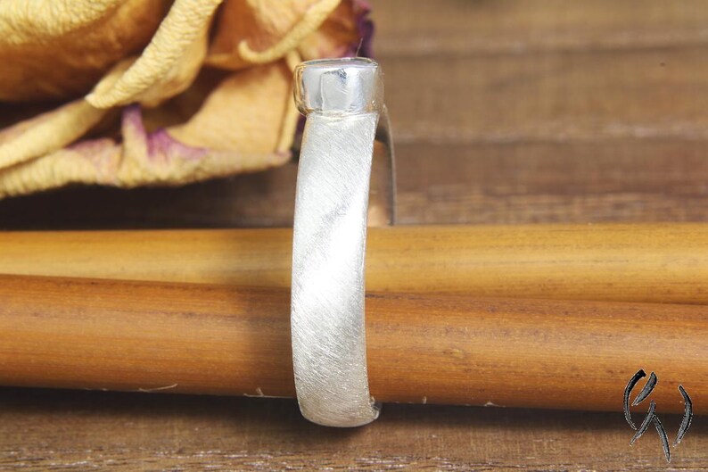 May include: A silver ring with a brushed finish and a round, flat top. The ring is resting on a wooden surface.