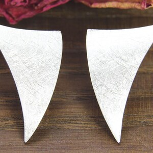 May include: A pair of silver earrings with a brushed metal finish. Each earring has a curved, abstract shape, resembling a stylized leaf or wing. The earrings are displayed on a wooden surface.
