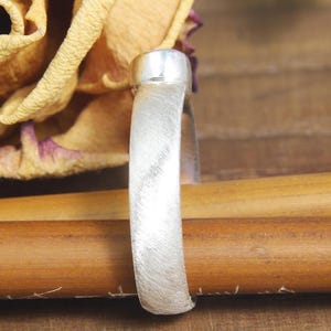 May include: A silver ring with a brushed finish. The ring is on a wooden surface.