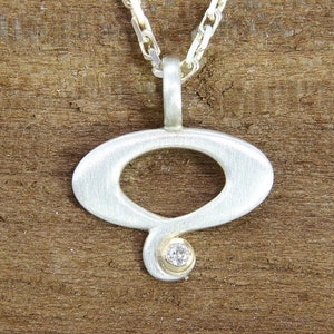 May include: A silver pendant necklace with a brushed finish, featuring a unique oval shape with a small diamond accent. The pendant hangs from a silver chain.