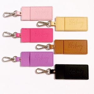 May include: Six keychains with different coloured leather card holders. Each keychain has a silver metal clasp and the text "Blusherry" embossed on the leather. The colours are pink, beige, brown, purple, and black.