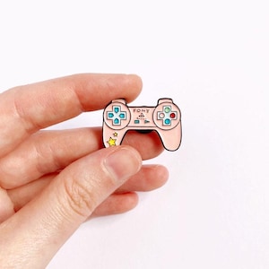 May include: A pink PlayStation controller enamel pin with a yellow star and the word "SONY" on it.