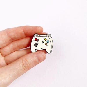 May include: White enamel pin of a video game controller with black outlines and colourful buttons.