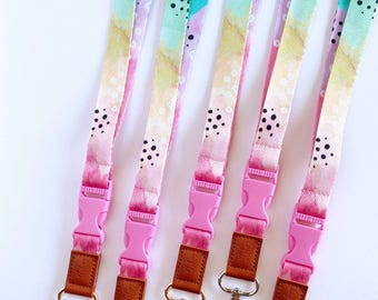 May include: Four colorful lanyards with a pink buckle, brown leather strap, and gold clip. The lanyards have a tie-dye pattern with black dots and a white background.