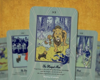 May include: A blue and green tarot card with the number XII and the words "The Hanged Man" printed on it. The card features an illustration of Dorothy from the Wizard of Oz, the Cowardly Lion, the Scarecrow, and Toto the dog. The text on the card reads "Experience is the only thing that brings knowledge, and the longer you are on earth the more experience you are sure to get."