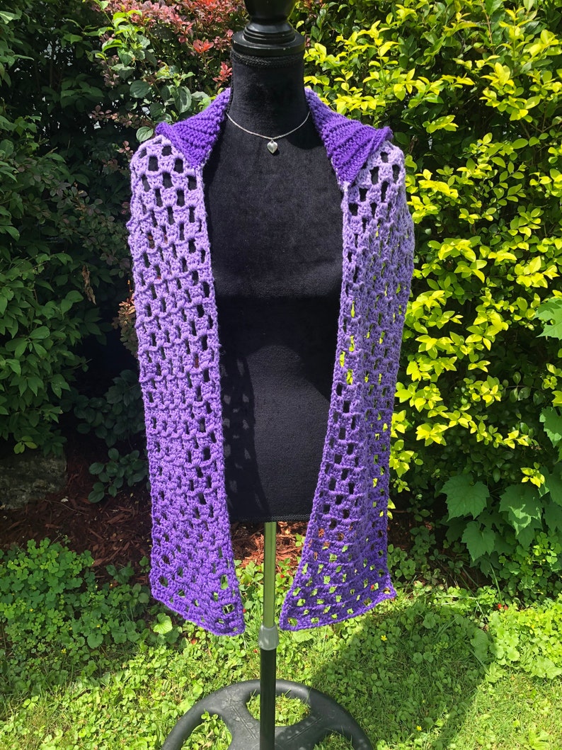 May include: A purple and lavender ombre crochet scarf with a hood. The scarf is displayed on a black mannequin.