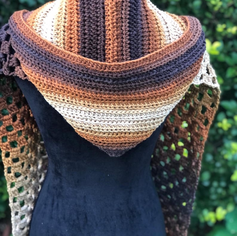 May include: A crocheted shawl with a hooded design in brown, beige, and tan colors. The shawl is made with a lacy stitch pattern and has a striped pattern on the hood.