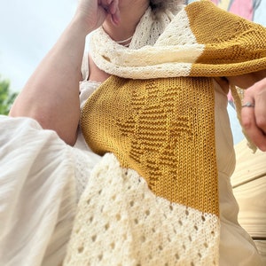 May include: A knitted scarf with a white and mustard yellow color scheme. The scarf has a lacy pattern on one side and a solid stitch on the other side.