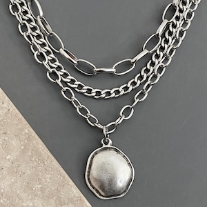 May include: A layered silver necklace with three chains. The top chain has large oval links, the middle chain is a curb chain, and the bottom chain has oval links and a pendant. The pendant is a textured, irregular shape.