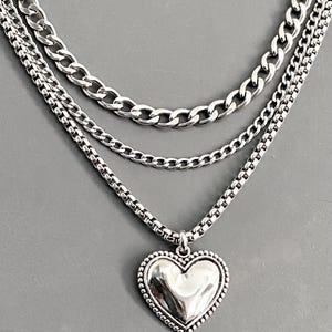 May include: A layered silver-tone necklace featuring three distinct chains of varying thicknesses. The bottom chain is a box chain that supports a central heart-shaped pendant with a smooth, polished surface and a beaded border detail.