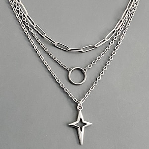 May include: A layered silver-tone necklace featuring three distinct chains. The top chain is a long oval link, the middle chain features a simple circular ring, and the bottom chain holds a four-pointed star-shaped pendant with a cutout center.