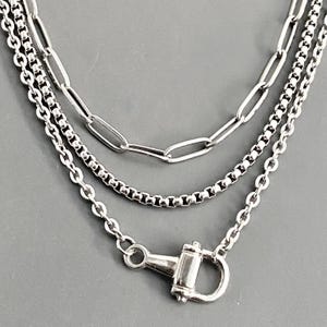 May include: A layered silver-toned necklace featuring three distinct chain styles: an elongated paperclip chain, a square box chain, and a standard cable chain. The necklace is secured with a prominent, polished silver-toned metal shackle clasp.
