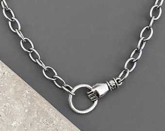 May include: A silver-toned metal chain necklace featuring a unique clasp design. The closure consists of a circular ring held by a sculpted hand motif, which connects to the main chain links. The metal has a polished finish with visible detailing.