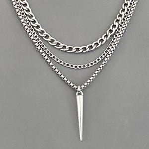 May include: A silver-toned layered necklace featuring three distinct chains of varying thicknesses. The outermost chain is a thick curb link, the middle is a delicate cable chain, and the innermost is a box chain with a pointed spike pendant.