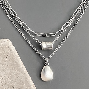 May include: A layered silver-tone necklace featuring three distinct chains. The top layer is a paperclip chain, the middle layer holds a cylindrical bead, and the bottom layer features a smooth, teardrop-shaped pendant with a polished finish.