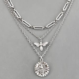 May include: A silver-toned layered necklace featuring three distinct chains. The top chain is a thick rectangular link, the middle chain holds a small bee charm, and the bottom chain features a round coin pendant embossed with a detailed bee design.