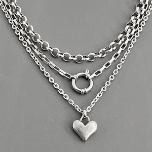 May include: A layered silver-toned necklace featuring three distinct chains. The top chain is a round link, the middle chain has rectangular links with a circular spring ring clasp, and the bottom chain holds a smooth, solid heart-shaped pendant.