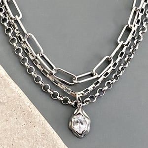 May include: A layered silver-tone necklace featuring three distinct chain styles: a large elongated oval link chain, a square box chain, and a round rolo chain. A single pendant with an irregular, molten-look frame holds a clear oval-cut gemstone.