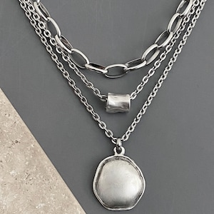 May include: A layered silver necklace with three strands. The top strand features large oval links, the middle has a cylindrical bead, and the bottom strand has a pendant with an irregular shape. The necklace is displayed on a gray surface.