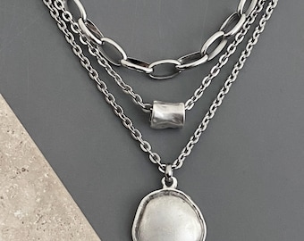 May include: A layered silver-tone necklace featuring three distinct chains. The top chain has large oval links, the middle chain includes a small cylindrical bead, and the bottom chain holds a large, irregular circular pendant with a matte finish.