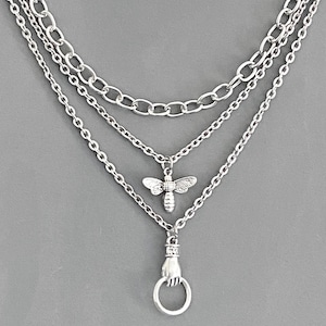 May include: A silver-toned layered necklace featuring three distinct chains. The middle chain displays a small bee charm, while the bottom chain features a pendant shaped like a hand holding a circular ring. The metal has a polished finish.