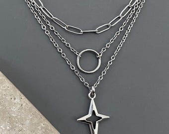 May include: A layered silver-tone necklace featuring three distinct chains. The top chain is a thick paperclip link, the middle chain holds a simple circular ring, and the bottom chain features a four-pointed star pendant with a hollow center.