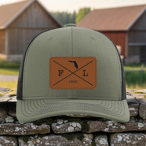 May include: A green and black trucker hat with a leather patch showing the outline of Florida, the letters "F" and "L", and the year "1845". The hat is on a stone wall, with barns and fields in the background.