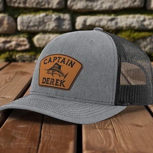 May include: Gray trucker hat with a black mesh back. A brown leather patch on the front displays a marlin graphic and the text "CAPTAIN DEREK". The hat sits on a wooden surface.