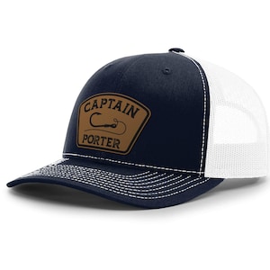 May include: Navy blue and white trucker hat with a curved brim. The hat features a brown leather patch with the words "CAPTAIN PORTER" and a fishing hook design. The hat has a mesh back and a snap closure.