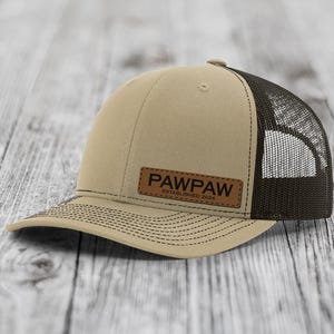 May include: Tan and black trucker hat with a curved brim. The hat features a tan front panel and a black mesh back. A brown leather patch on the front displays the word "PAWPAW" and the text "ESTABLISHED 2024".