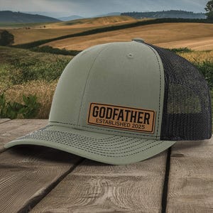 May include: A green and black trucker hat with the word "GODFATHER" and "ESTABLISHED 2025" printed on a brown patch. The hat is sitting on a wooden surface with a scenic background.