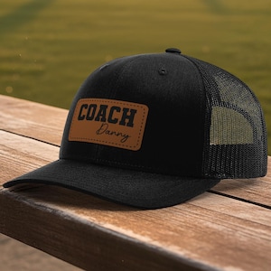May include: Black trucker hat with a mesh back and a curved brim. The hat features a brown leather patch with the words "COACH Danny" in bold, black lettering. The hat is sitting on a wooden surface.