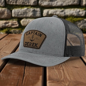 May include: Gray trucker hat with a black mesh back. The hat features a brown leather patch with the words "CAPTAIN DEREK" and an anchor graphic. The hat is sitting on a wooden surface.