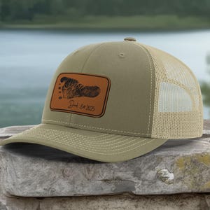 May include: Olive green trucker hat with a tan mesh back. A rectangular brown leather patch features a footprint design and the text "Dad Est. 2025". The hat has a curved brim and a button on top.