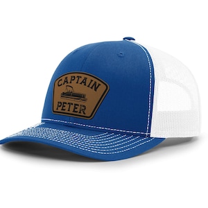 May include: A blue and white trucker hat with a curved brim. The front panel is blue, and the back is white mesh. A brown patch with the words "CAPTAIN PETER" and a boat graphic is on the front.