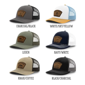 May include: A collection of six trucker hats in various color combinations, each featuring a leather patch with nautical-themed designs and the word "CAPTAIN" or "FIRST MATE" along with a name. The hats include charcoal/black, white/sky/yellow, loden, navy/white, khaki/coffee, and black/charcoal.