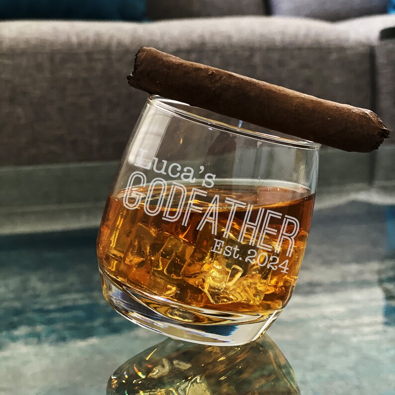 May include: A clear glass with a brown liquid and ice cubes. The glass has the text "Luca's Godfather Est. 2024" etched on the side. A brown cigar is resting on the rim of the glass.