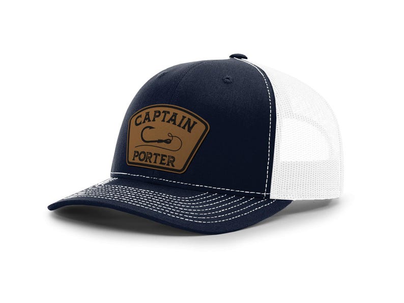 May include: Navy blue and white trucker hat with a curved brim. The hat features a brown leather patch with the words "CAPTAIN PORTER" and a fishing hook design. The hat has a mesh back and a snap closure.
