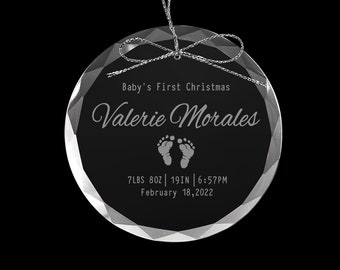 May include: A clear glass Christmas ornament with a silver ribbon bow. The ornament is engraved with "Baby's First Christmas", "Valerie Morales", a pair of baby footprints, "7 LBS 8OZ | 19IN | 6:57PM", and "February 18, 2022".
