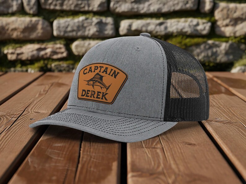 May include: Gray trucker hat with a black mesh back. A brown leather patch on the front displays a marlin graphic and the text "CAPTAIN DEREK". The hat sits on a wooden surface.